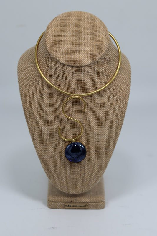 Handmade Kenyan brass choker necklace featuring a rigid collar with an artistic S-curve drop holding a deep blue circular stone, displayed on a burlap jewelry bust
