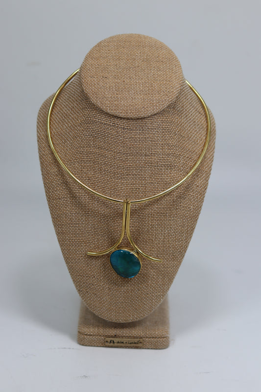 Handmade Kenyan brass choker necklace displayed on a beige burlap bust, featuring a smooth gold-tone collar wire and an architectural drop pendant with parallel vertical wires flaring outward above a polished teal-blue stone.
