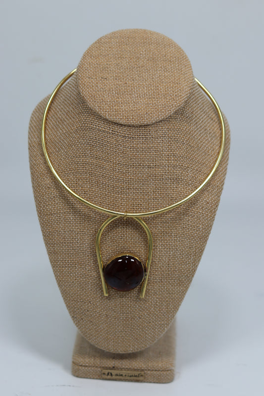 Handmade Kenyan brass choker necklace displayed on a beige burlap bust, featuring a smooth gold-tone collar wire and an inverted U-shaped drop pendant holding a polished glossy dark red stone.