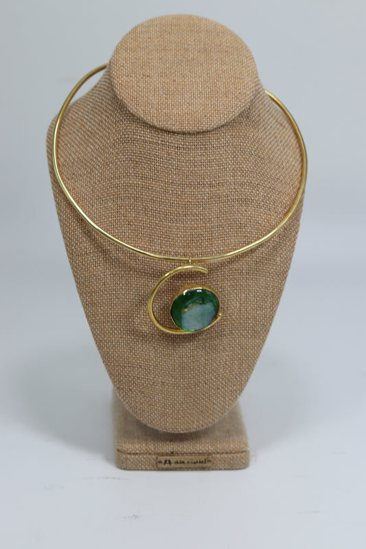 Handmade Kenyan brass choker necklace displayed on a beige burlap bust, featuring a smooth gold-tone collar wire and a circular curved brass pendant holding a polished green stone.