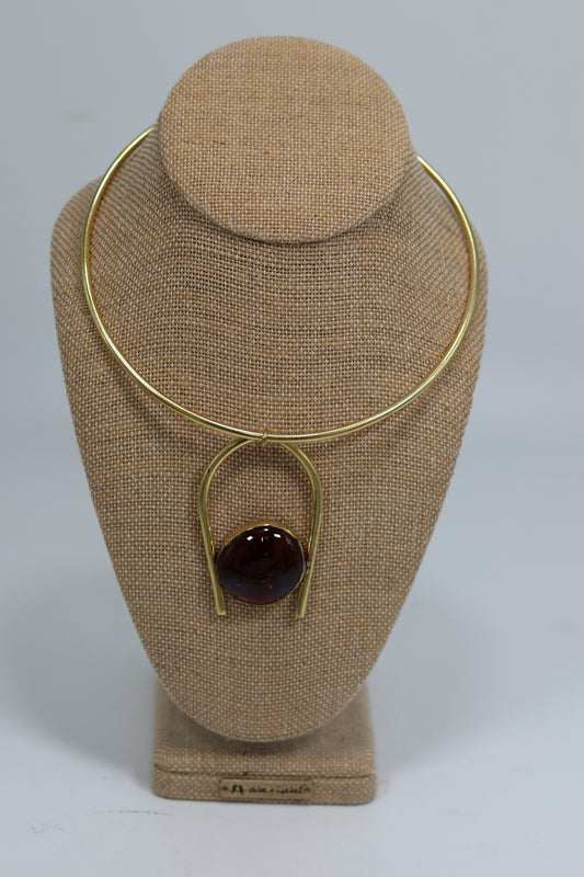 Handmade Kenyan brass choker necklace displayed on a beige burlap bust, featuring a sleek gold-tone wire neck ring and a bold U-shaped drop pendant with a glossy dark red stone.