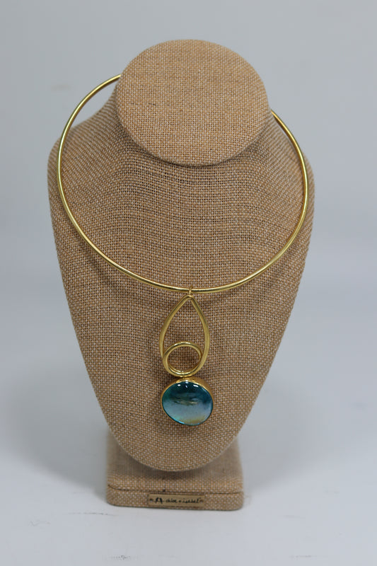 Handmade Kenyan brass choker necklace displayed on a beige burlap bust, featuring a smooth gold-tone collar wire and an geometric drop pendant with a teardrop loop, a small ring, and a polished blue-green stone.