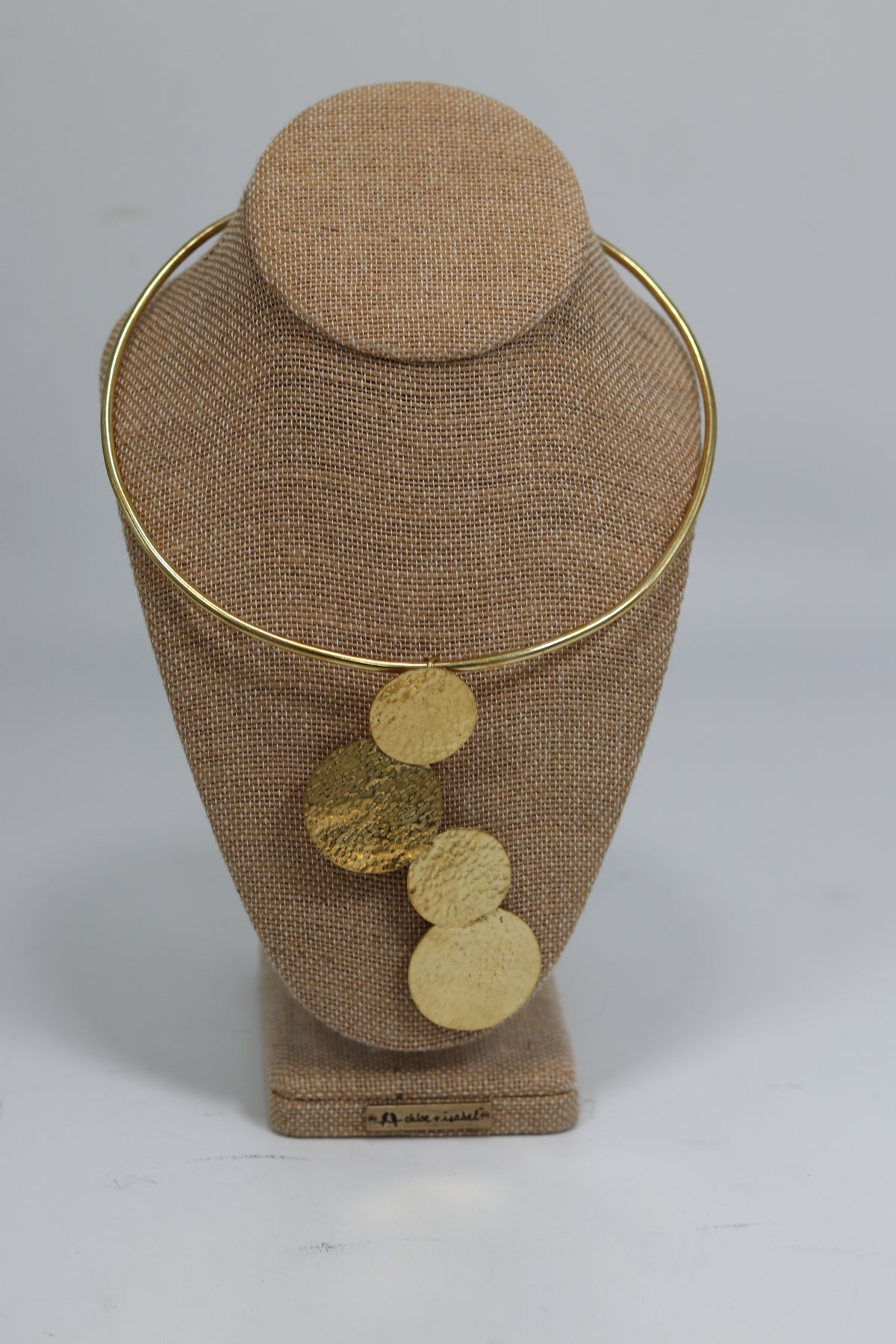 Hand-hammered Kenyan brass choker necklace displayed on a beige burlap bust, featuring a smooth gold-tone neck collar with four asymmetrical cascading textured round brass discs.